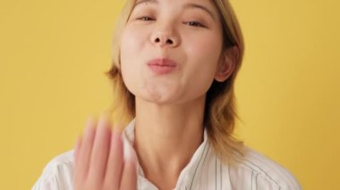 Yakın plan, gülümseyen genç bir kadın stüdyodaki sarı arka planda izole edilmiş kameraya hava öpücüğü gönderiyor.