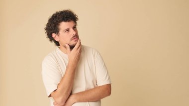 Düşünceli, kıvırcık saçlı, bej tişörtlü, elleri katlı, stüdyoda bej arka planda karar veremeyen biri.
