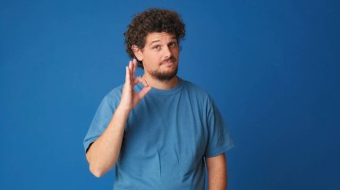 Mavi tişörtlü, kıvırcık saçlı, gülümseyen adam kameraya bakıyor ve stüdyonun mavi arka planında izole edilmiş bir hareket sergiliyor.