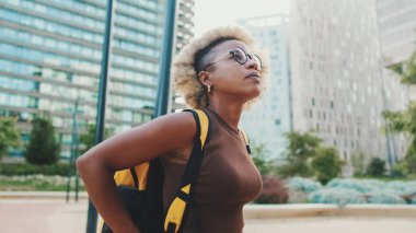 Gözlüklü, kahverengi elbiseli, sırtında sırt çantasıyla genç bir kadın modern binalarda sokakta yürüyor.