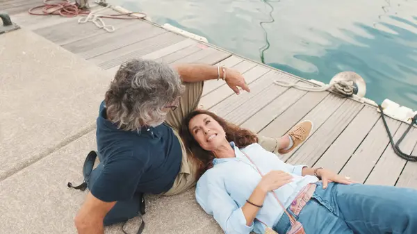 Happy middle aged couple, retired elderly couple enjoy joint outdoor recreation, relax and chat lying on pier on yacht background