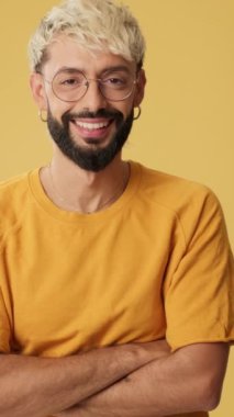 Genç adam kollarını kavuşturup gülüyor. Stüdyoda sarı arka planda izole edilmiş kameraya bakıyor.