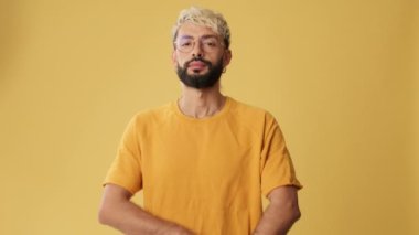 Genç adam kollarını kavuşturuyor ve gülümsüyor. Stüdyoda sarı arka planda izole edilmiş kameraya bakıyor.