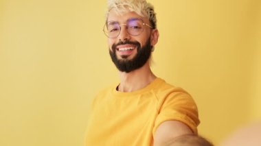 Yakın plan, adam kameraya bakıyor selfie çekiyor, sarı arkaplanda zafer işaretini gösteriyor.