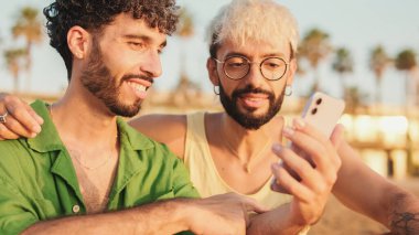 Sahilde oturan, gülümseyen, konuşan ve cep telefonu kullanan genç eşcinsel çiftin yakın çekimi.