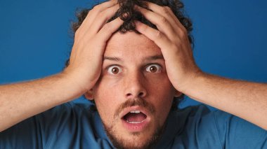 Yakından, mavi tişörtlü, kıvırcık saçlı, büyük gözlü, kameraya bakan, ellerini kaldıran ve stüdyodaki mavi arka planda izole edilmiş iyi haberden memnun olan adam.