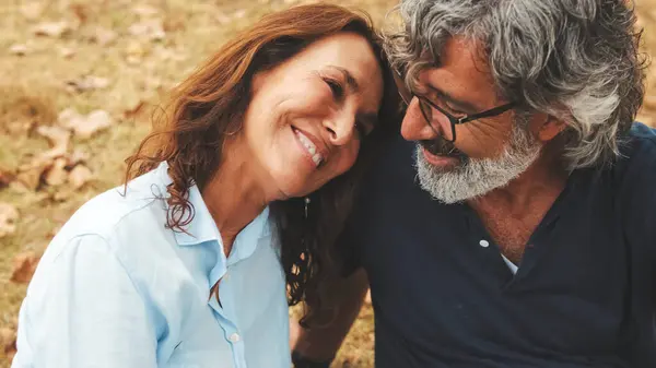 Mutlu emekli çiftin sonbaharda parkta çimlerin üzerinde otururken birlikte gülümseyerek ve rahatlayarak vakit geçirdiği yakın plan.