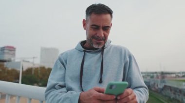 Spor giyim sektöründe orta yaşlı bir adam köprüde dururken cep telefonu kullanıyor.