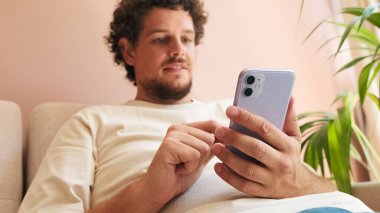 Kıvırcık saçlı ve sakallı genç adam, rahat oturma odasında koltukta oturuyor, cep telefonu kullanıyor.