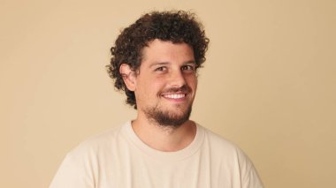 Kıvırcık saçlı, bej tişörtlü mutlu adam stüdyoda izole edilmiş kameraya bakıp gülüyor.