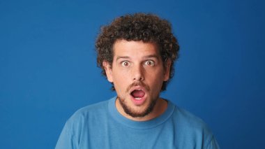 Yakından, mavi tişörtlü, şaşkın adam kameraya büyük gözlerle bakıyor, stüdyonun mavi arka planında izole edilmiş.