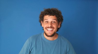 Yaklaş, mavi tişörtlü, kıvırcık saçlı, kameraya gülümseyen, stüdyonun mavi arka planında soyutlanmış adam.