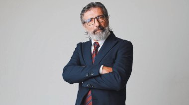 Gözlüklü, gri saçlı orta yaşlı bir işadamı kollarını kavuşturup kameraya bakıyor. Stüdyonun gri arka planında tek başına.