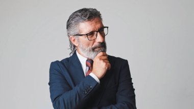 Orta yaşlı gözlüklü, gri saçlı, el destekli çenesi olan, stüdyonun gri arka planında soyutlanmış bir iş adamı.