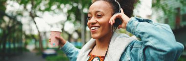 Kot ceketli, gülen Afrikalı bir kız. Üstünde ulusal modelle sokakta yürüyor. Kulaklıkla müzik dinliyor ve dans ediyor.