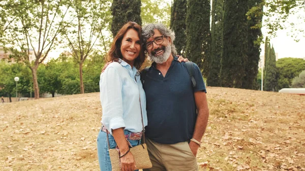 Smiling older married couple looking at the camera while standing in the park hugging each other