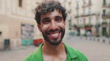 Yakından, sakallı, yeşil tişörtlü, kameraya bakarken gülümseyen esmer adam.