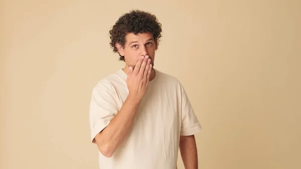 Guy with curly hair dressed in beige t-shirt looking at camera and embarrassedly covering his mouth with his hand, isolated on beige background in studio
