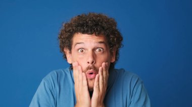 Yaklaş, mavi tişörtlü, şaşkın adam kameraya büyük gözlerle bakıyor, ellerini yüzüne kaldırıyor, vay, stüdyonun mavi arka planında izole edilmiş.