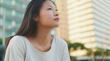 Düşünceli güzel esmer kız, günlük giysiler içinde bankta oturuyor ve başka tarafa bakıyor.