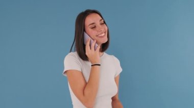 Cep telefonuyla konuşan genç bir kadın stüdyoda mavi arka planda izole edilmiş.