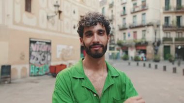 Sakallı, yeşil tişörtlü yakışıklı esmer adam kameraya bakarken kollarını kavuşturuyor.