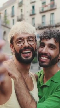 Dikey video, gay çift ayakta sarılıyor ve cep telefonundan video görüşmesi yapıyor, selamlaşırken el sallıyorlar.