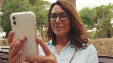 Güzel, gülümseyen, gözlüklü, parkta otururken telefonunu kullanan yaşlı bir kadının yakın çekimi.