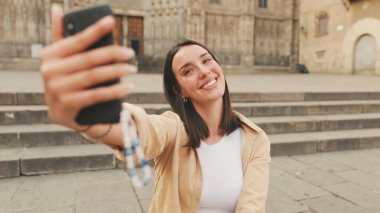 Gezgin kız, cep telefonuyla selfie çekiyor, eski bir Avrupa şehrinin tarihi bölümünde eski bir binanın merdivenlerinde otururken zafer işareti gösteriyor.