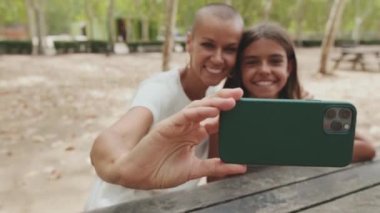 Parkta oturup cep telefonuyla selfie çeken mutlu anne ve kız.