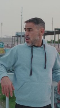 Dikey video, spor giyimli orta yaşlı adam köprüde koşarken nefesini toparlıyor.