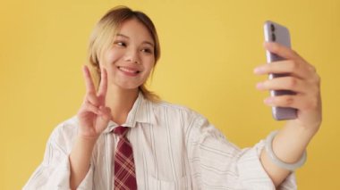 Cep telefonuyla selfie çeken genç bir kadın, zafer jesti, sarı arka planda izole edilmiş.