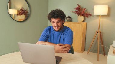 Kıvırcık saçlı ve sakallı olumlu bir genç arkadaşlarıyla uzaktan iletişim kurmak için bilgisayarını kullanıyor.