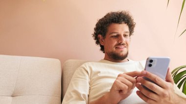 Kıvırcık saçlı, sakallı, rahat oturma odasında koltukta oturan, cep telefonu kullanan pozitif bir genç.