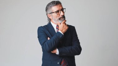 Orta yaşlı gözlüklü, gri saçlı, el destekli çenesi olan, stüdyonun gri arka planında soyutlanmış bir iş adamı.