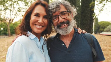 Yaklaş, gülümseyen yaşlı evli çift parkta birbirlerine sarılırken kameraya bakıyor.