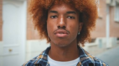 Afrikalı saçlı, mavi gömlekli, gülümseyen adam sokakta dikilirken kameraya bakıyor.