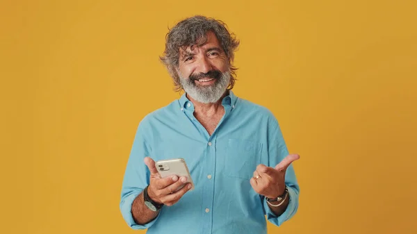 Elderly grey-haired bearded man wears a blue shirt, looks enthusiastically at a mobile phone, points to the phone while looking at the camera, isolated on an orange background in the studio