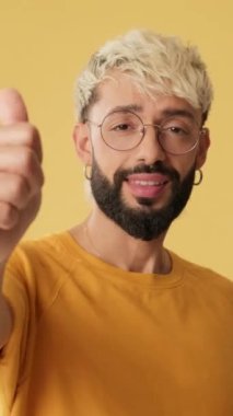 Yaklaş, başparmak hareketi yapan adam, stüdyoda sarı arkaplanda izole edilmiş kameraya bakıyor.