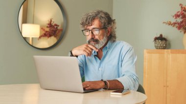 Beyaz saçlı, gözlüklü, oturma odasında dizüstü bilgisayar kullanan yaşlı bir adam.