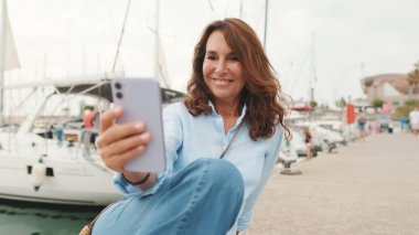 Güzel, zayıf, orta yaşlı bir kadın yat arka planında limanda oturuyor ve selfie cep telefonu yapıyor.