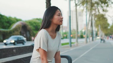 Güzel, mutlu esmer kız günlük giysiler içinde dışarıda bankta oturuyor.