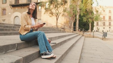 Eski bir Avrupa şehrinin tarihi bölümünde eski bir binanın merdivenlerinde oturan gezgin kız cep telefonu kullanıyor.