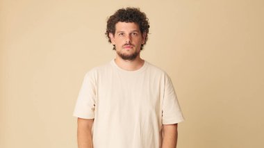 Kıvırcık saçlı, bej tişörtlü, büyük gözlü, stüdyoda bej arka planda dururken kameraya bakan harika bir adam.