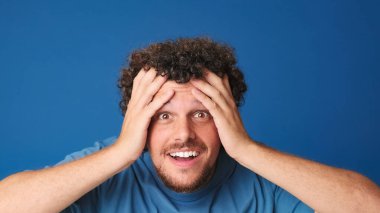 Yaklaş, mavi tişörtlü, şaşkın adam kameraya büyük gözlerle bakıyor, ellerini yüzüne kaldırıyor, vay, stüdyonun mavi arka planında izole edilmiş.