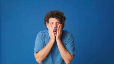 Mavi tişörtlü, kıvırcık saçlı bir adam kameraya büyük gözleriyle bakıyor ve ellerini kaldırıyor. Stüdyonun mavi arka planındaki iyi habere seviniyor.