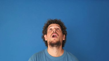 Kıvırcık saçlı, mavi tişörtlü, parmaklarıyla yer gösteren, göz reklamı yapan ya da stüdyoda mavi arka planda duyuru yapan, gülümseyen bir adam.