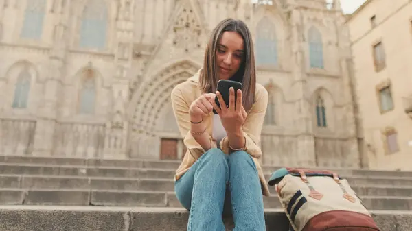 Happy traveler girl using mobile phone while sitting on the steps of an old building in the historic part of an old European city