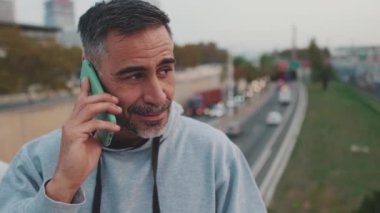 Yakın plan, spor kıyafetli orta yaşlı adam köprüde dururken cep telefonuyla konuşuyor.