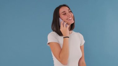 Cep telefonuyla konuşan gülen genç bir kadın stüdyoda mavi arka planda izole edilmiş.